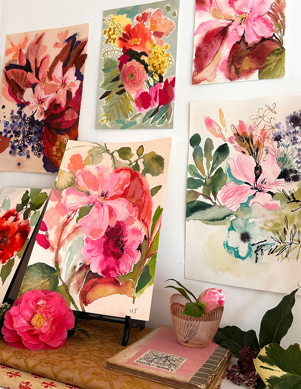 Collection of floral paintings on a wall with a notebook and pink flowers in the foreground.