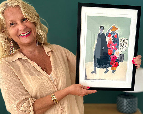Woman holding a framed artwork of two people with flowers against a teal wall.