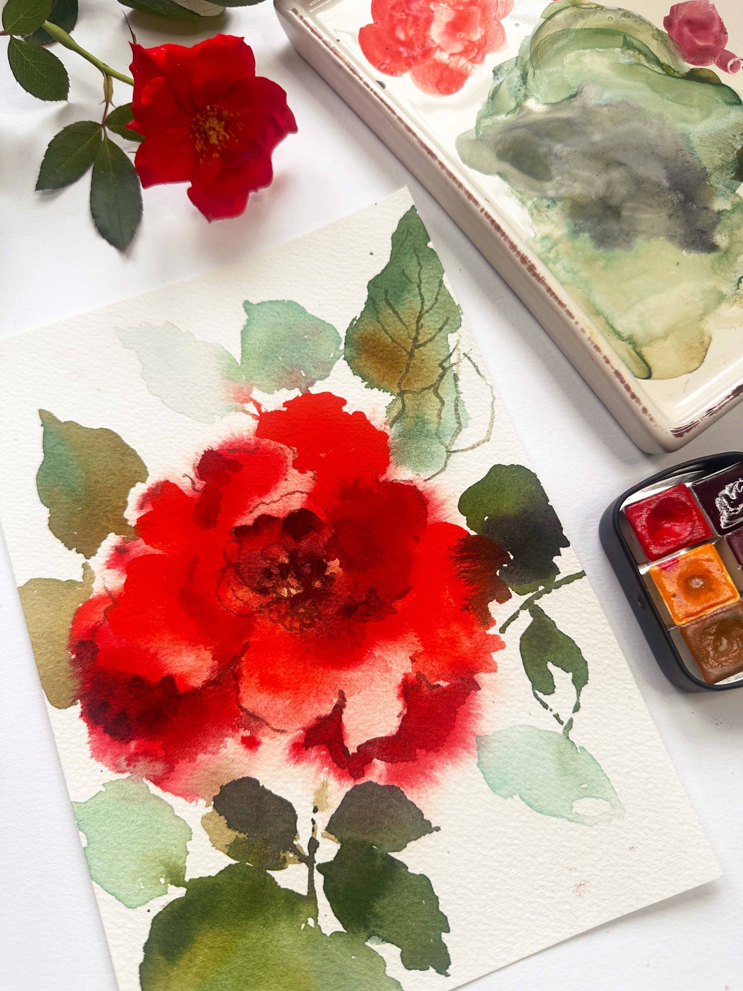 Watercolor painting of a red flower on a white surface with watercolor pans and a paintbrush.